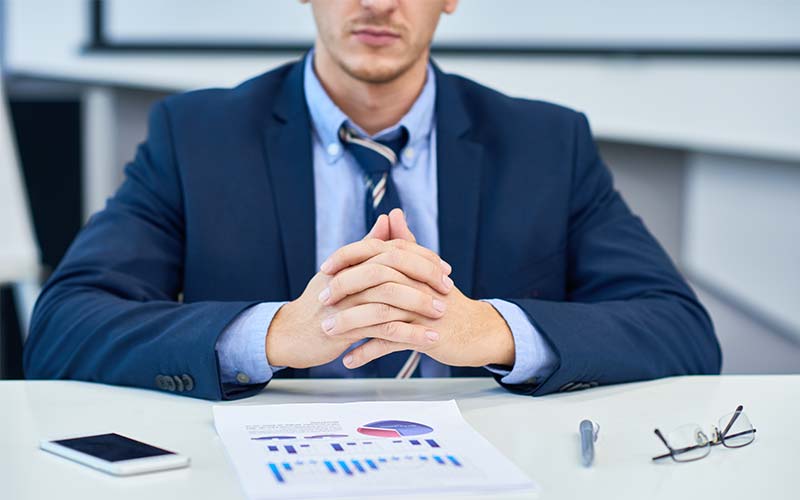 Man in a suit seated at a desk with a printed chart, a phone and reading glasses