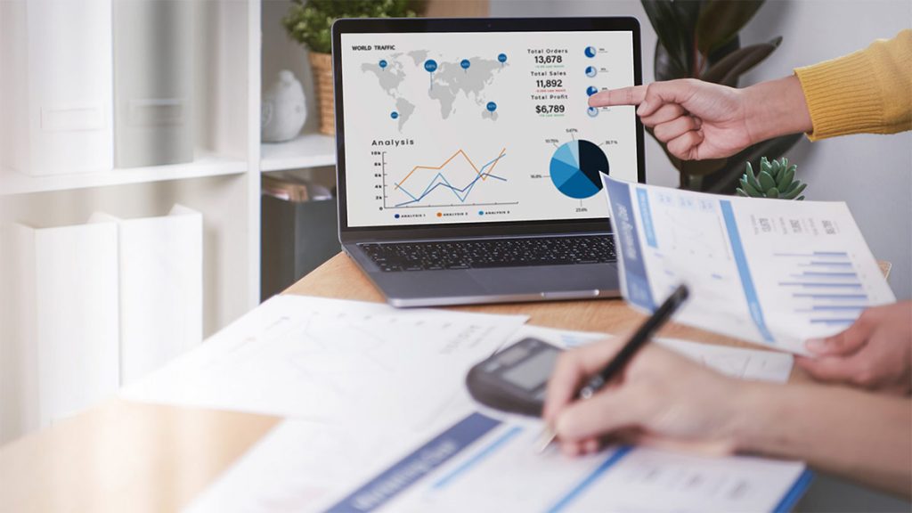 Laptop showing charts beside printed financial reports being reviewed with a pen