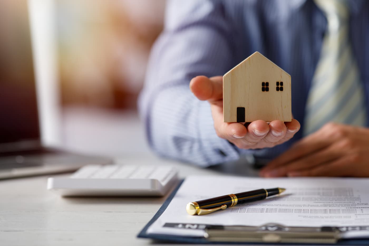 Hand holding a small wooden model house above a desk with a clipboard and pen